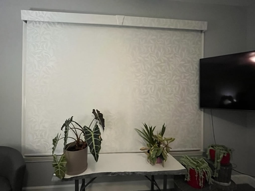 A thick fabric roller shade installed on a large window with a TV next to it hanging on the wall.